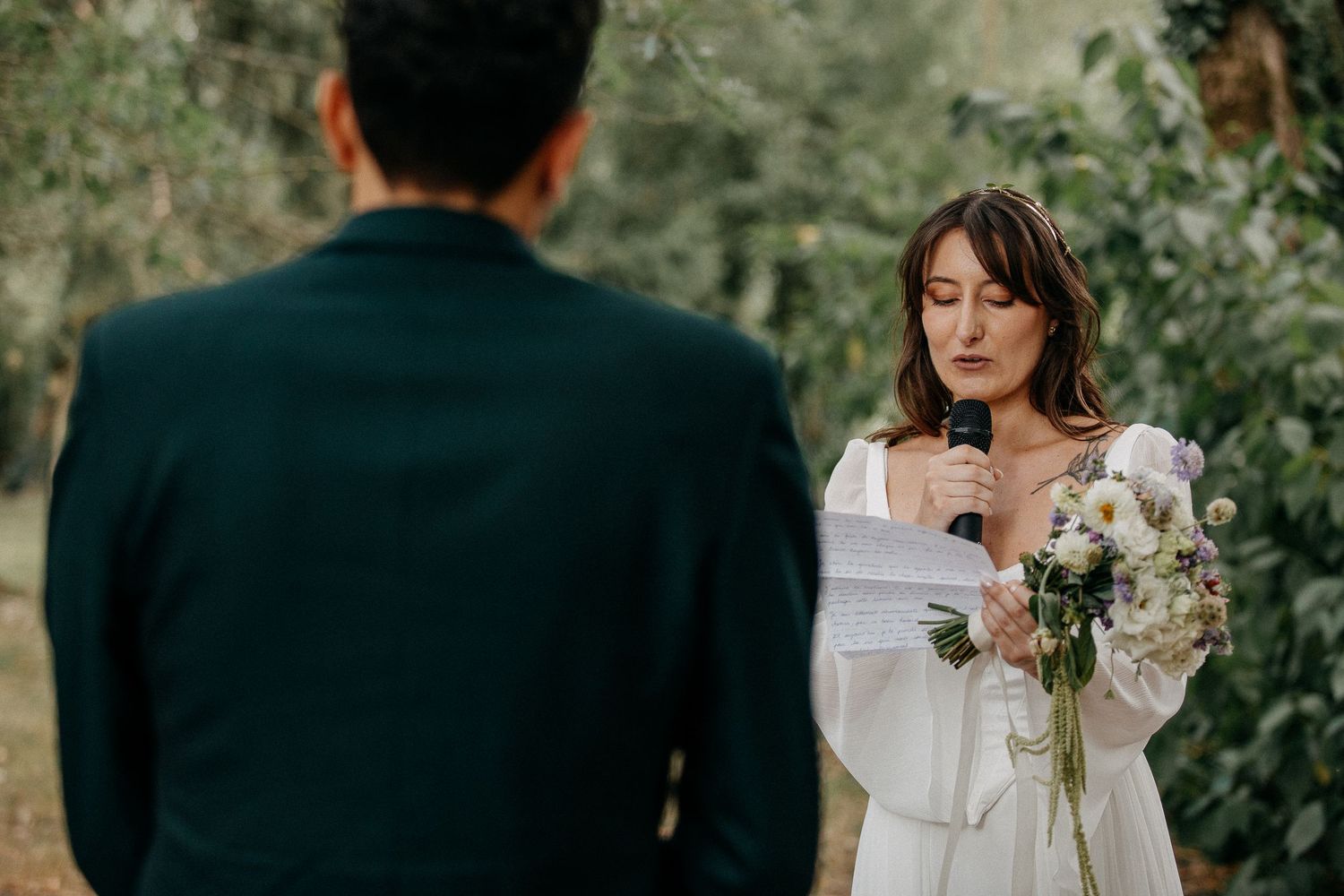 Bride in white dress reads vows holding bouquet at outdoor wedding ceremony.