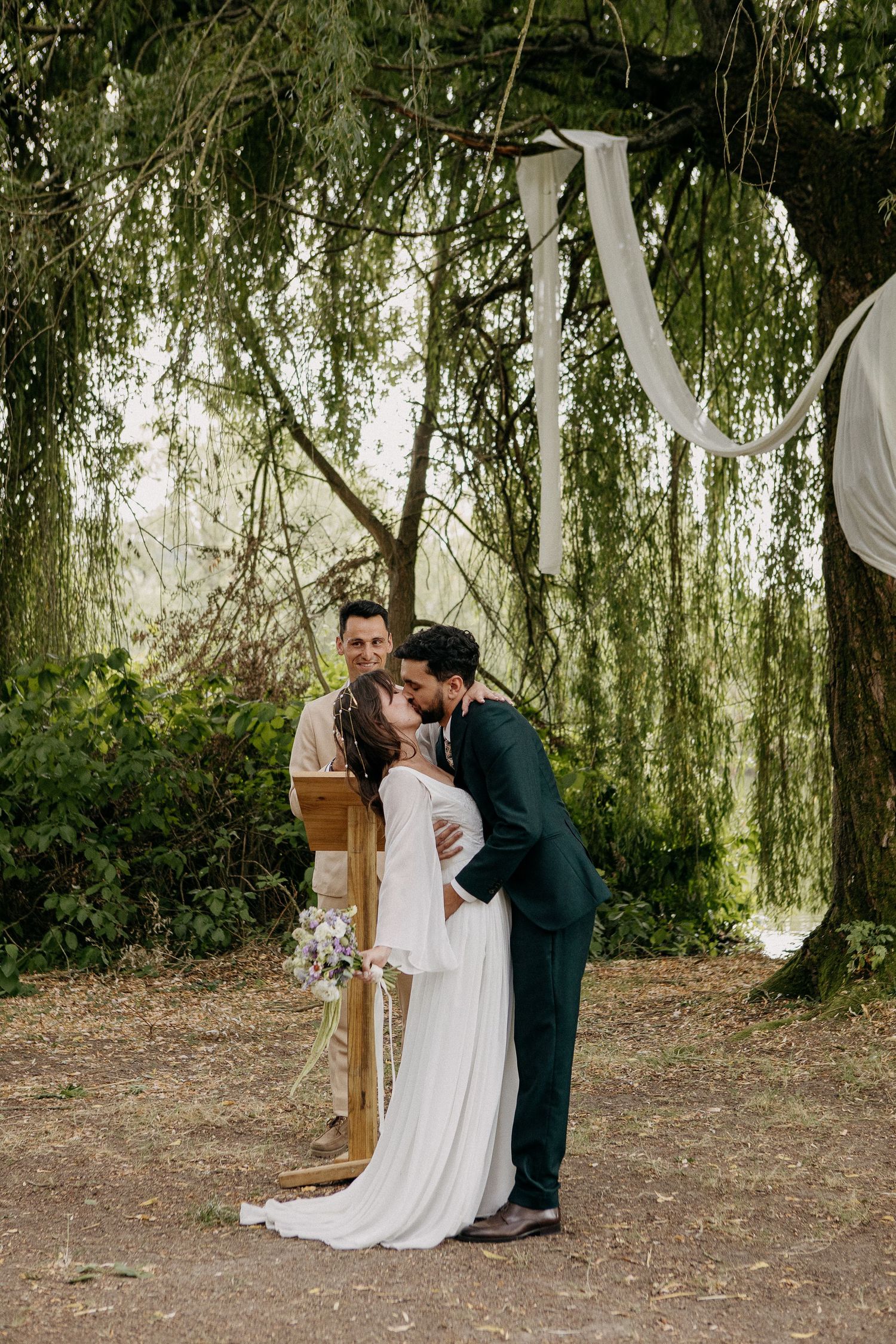 Bride and groom share first kiss under willow tree at outdoor wedding ceremony with officiant smiling behind them.