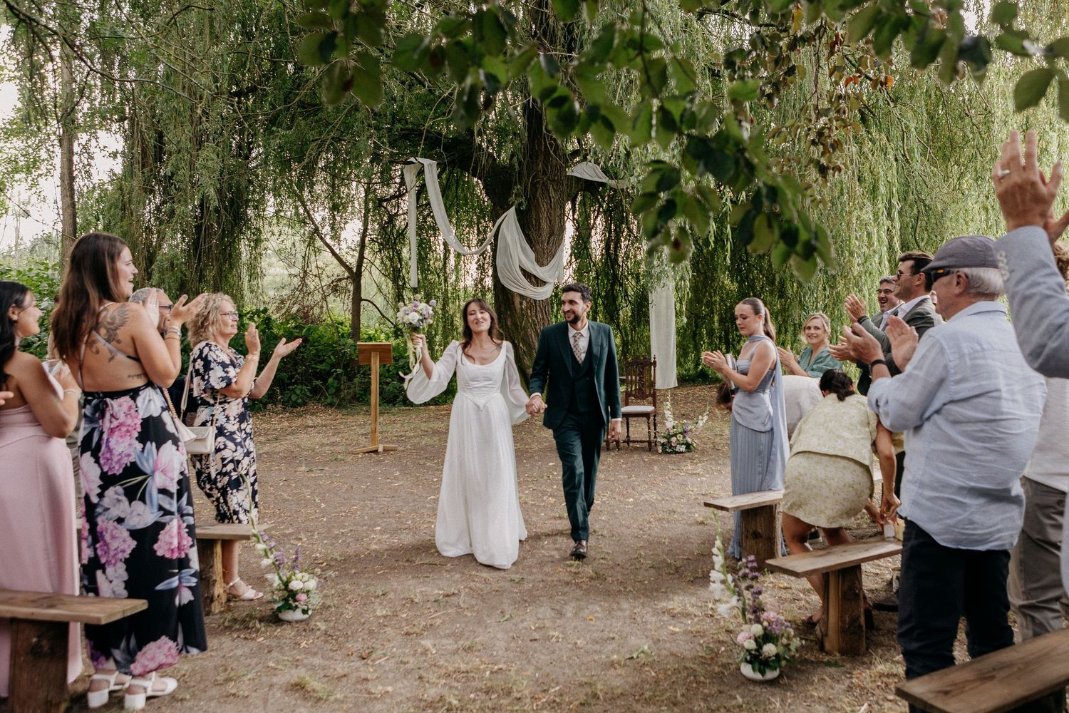 Newlywed couple walks down outdoor aisle under willow tree, guests applauding at rustic garden wedding ceremony.
