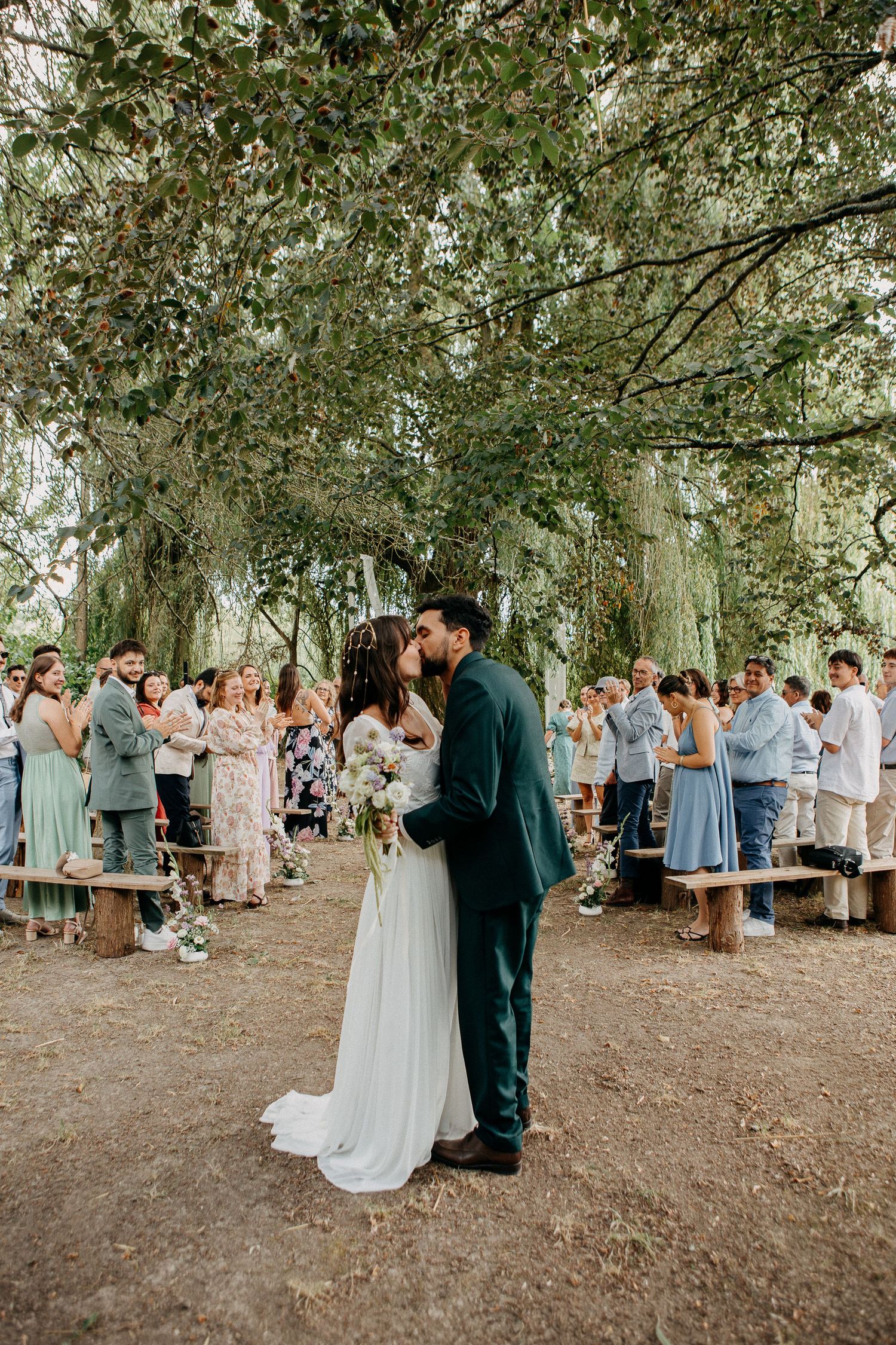 A bride and groom share their first kiss under a canopy of trees surrounded by wedding guests at an outdoor ceremony.