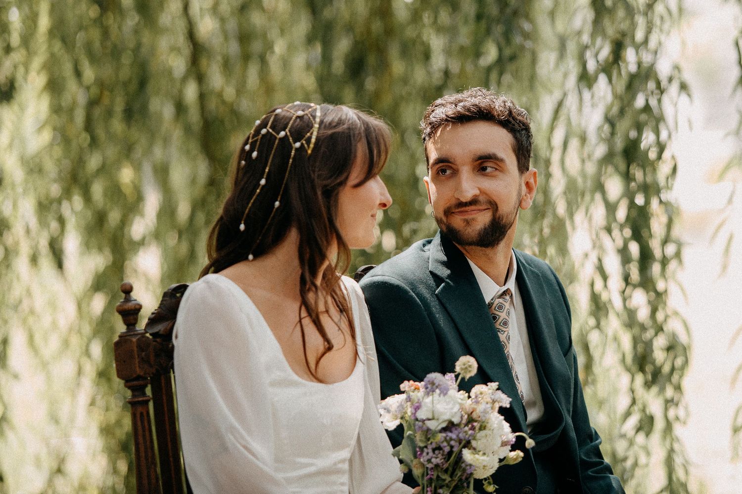 Bride and groom sitting together under a weeping willow tree, bride holding a bouquet of purple and white flowers.