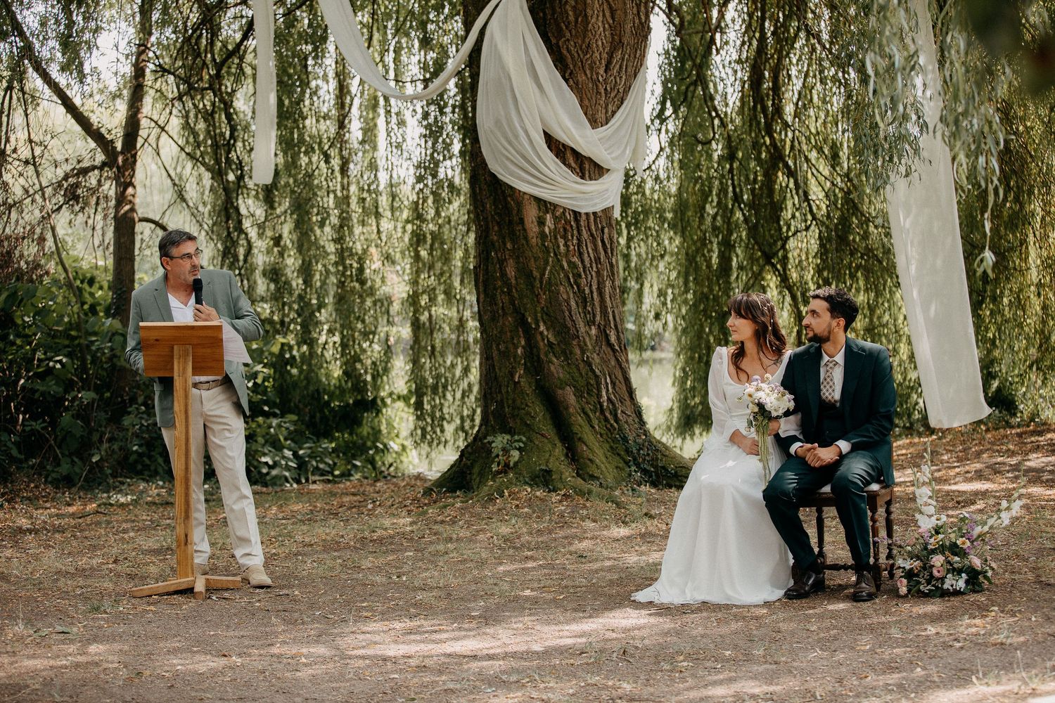 Outdoor woodland wedding ceremony with couple seated before officiant at wooden podium, draped fabric on tree.