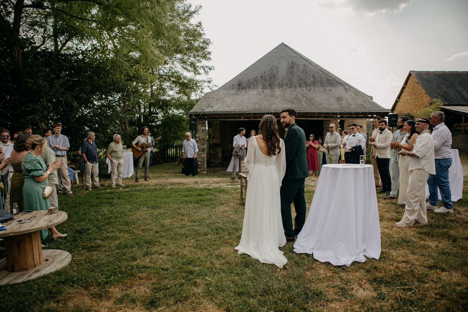 Outdoor wedding ceremony with bride and groom at altar, guests gathered on lawn beside rustic stone barn.