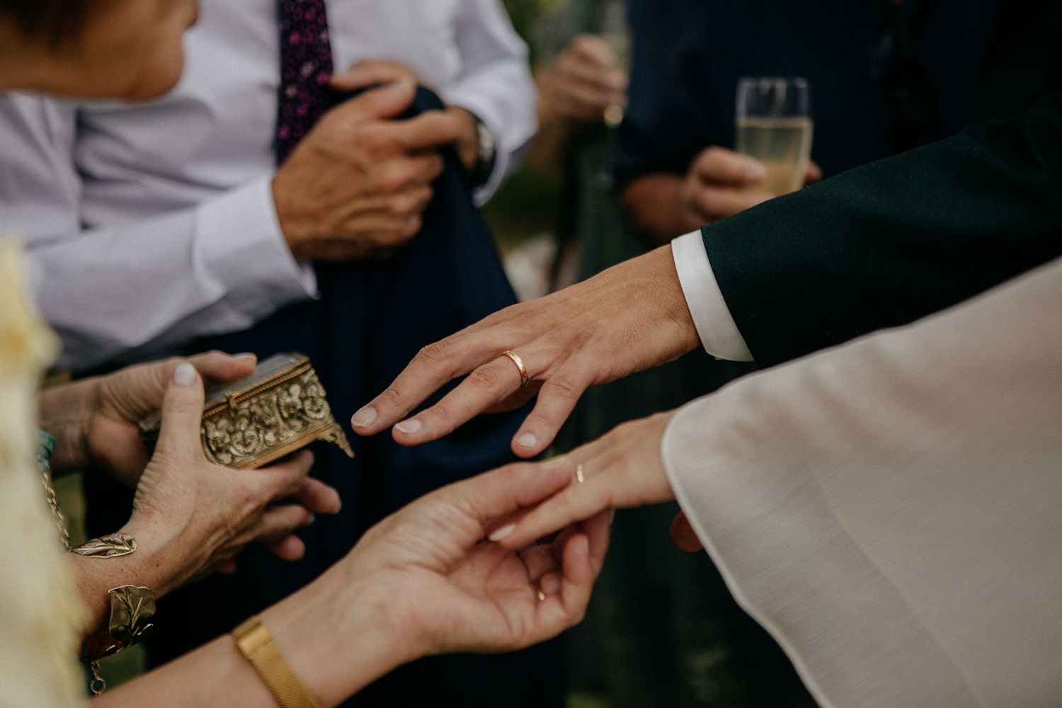 Close-up of wedding ring exchange ceremony with guests holding a decorative gold ring box during the special moment.