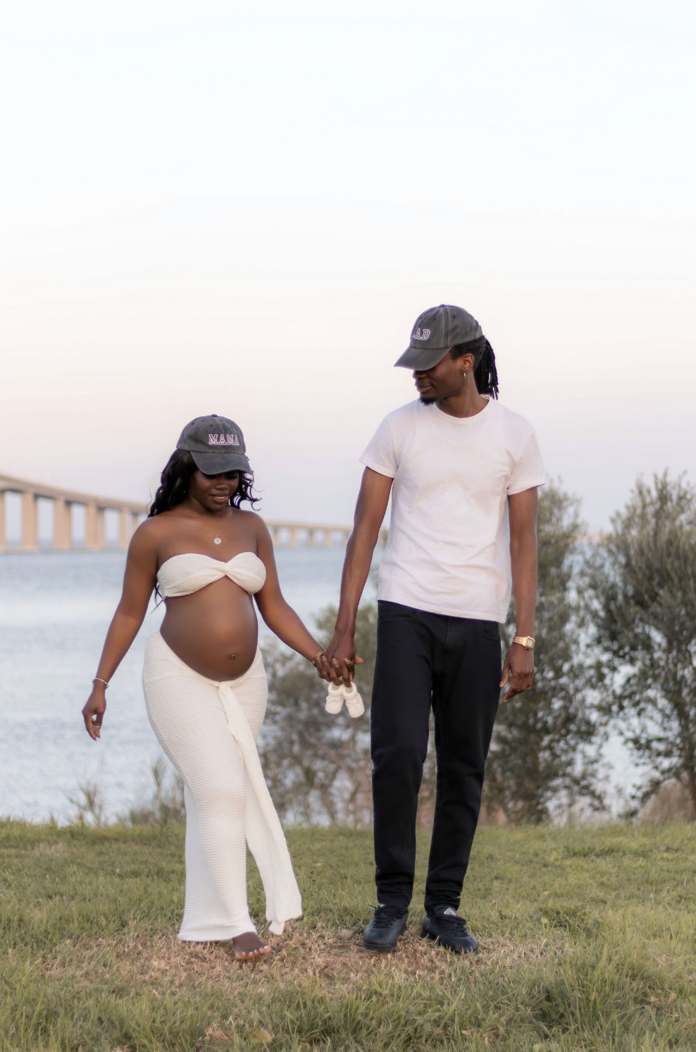 Pregnant couple holding hands outdoors near a bridge at sunset, woman in white dress showing baby bump, man in white tee.