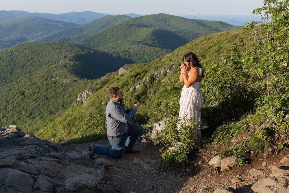 Man proposes on one knee to woman on rocky mountain overlook with lush green valley views at sunset.