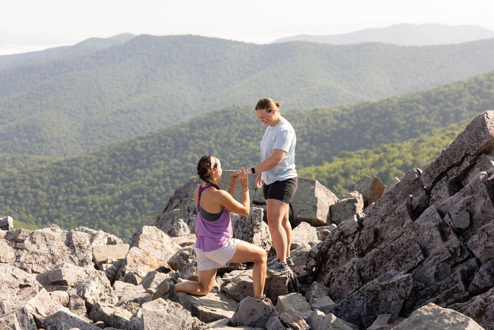 Person proposing on one knee atop a rocky mountain summit with scenic green forested hills in the background.