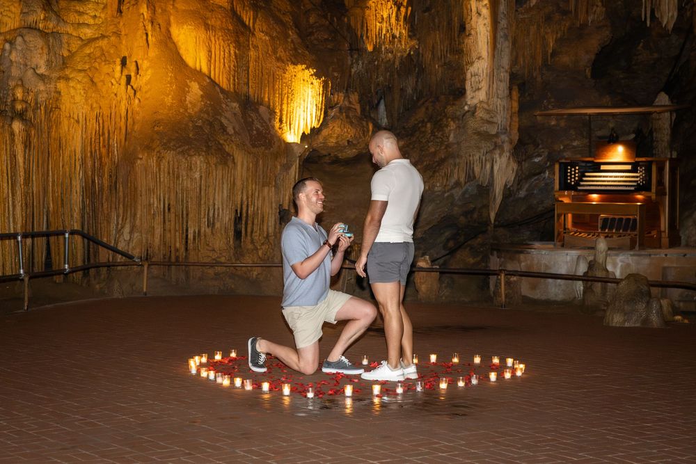 Man proposes to partner on one knee inside a cave decorated with candles arranged in a heart shape.
