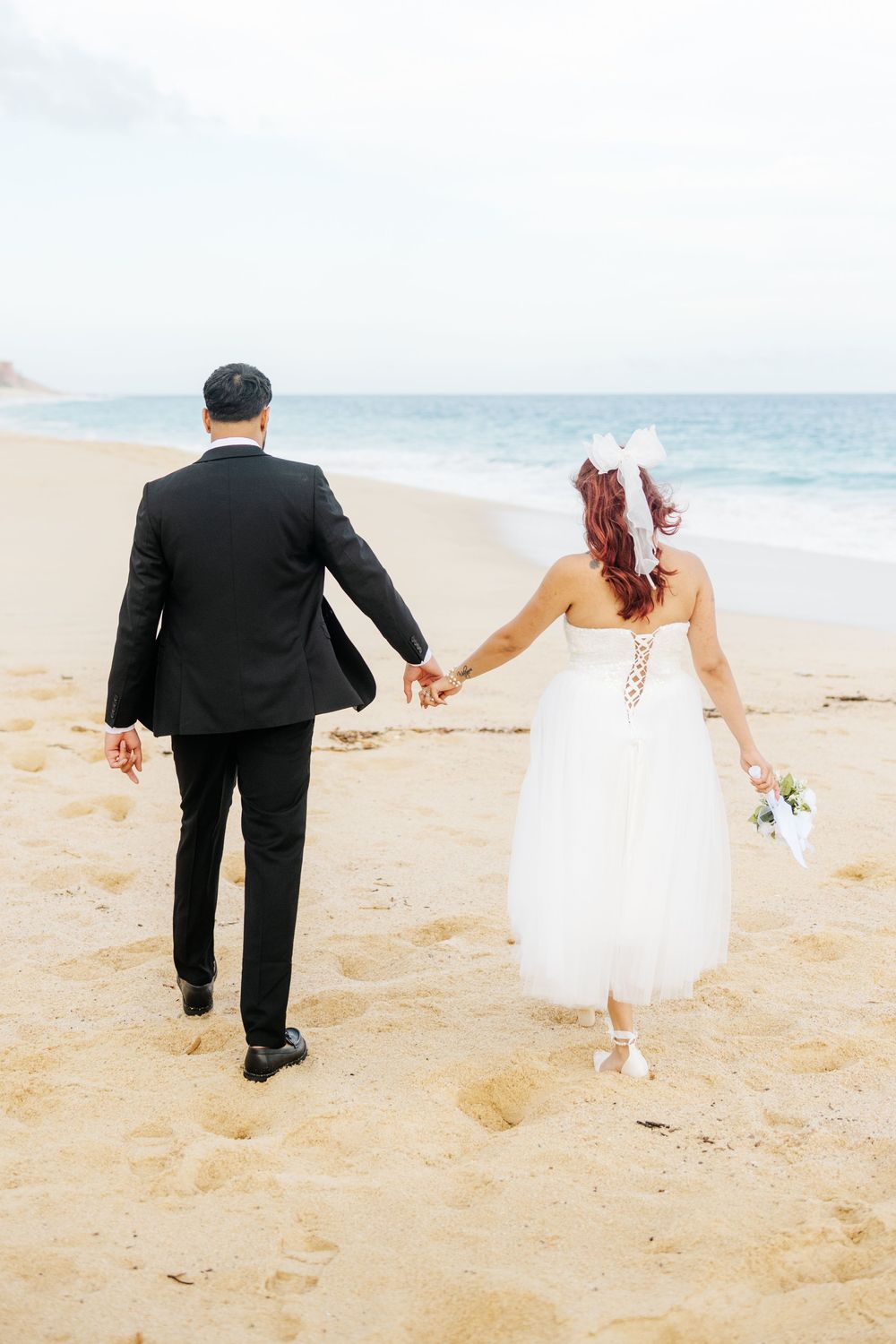 Newlywed couple holding hands walking on sandy beach, bride in white gown with bouquet, groom in black suit.