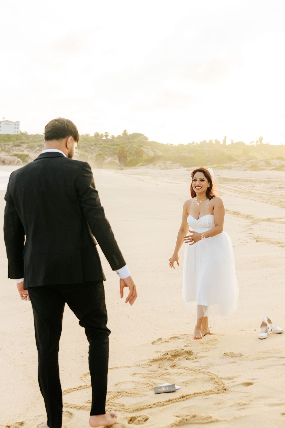 Bride in white dress laughs barefoot on sandy beach as groom in black suit approaches during golden hour.