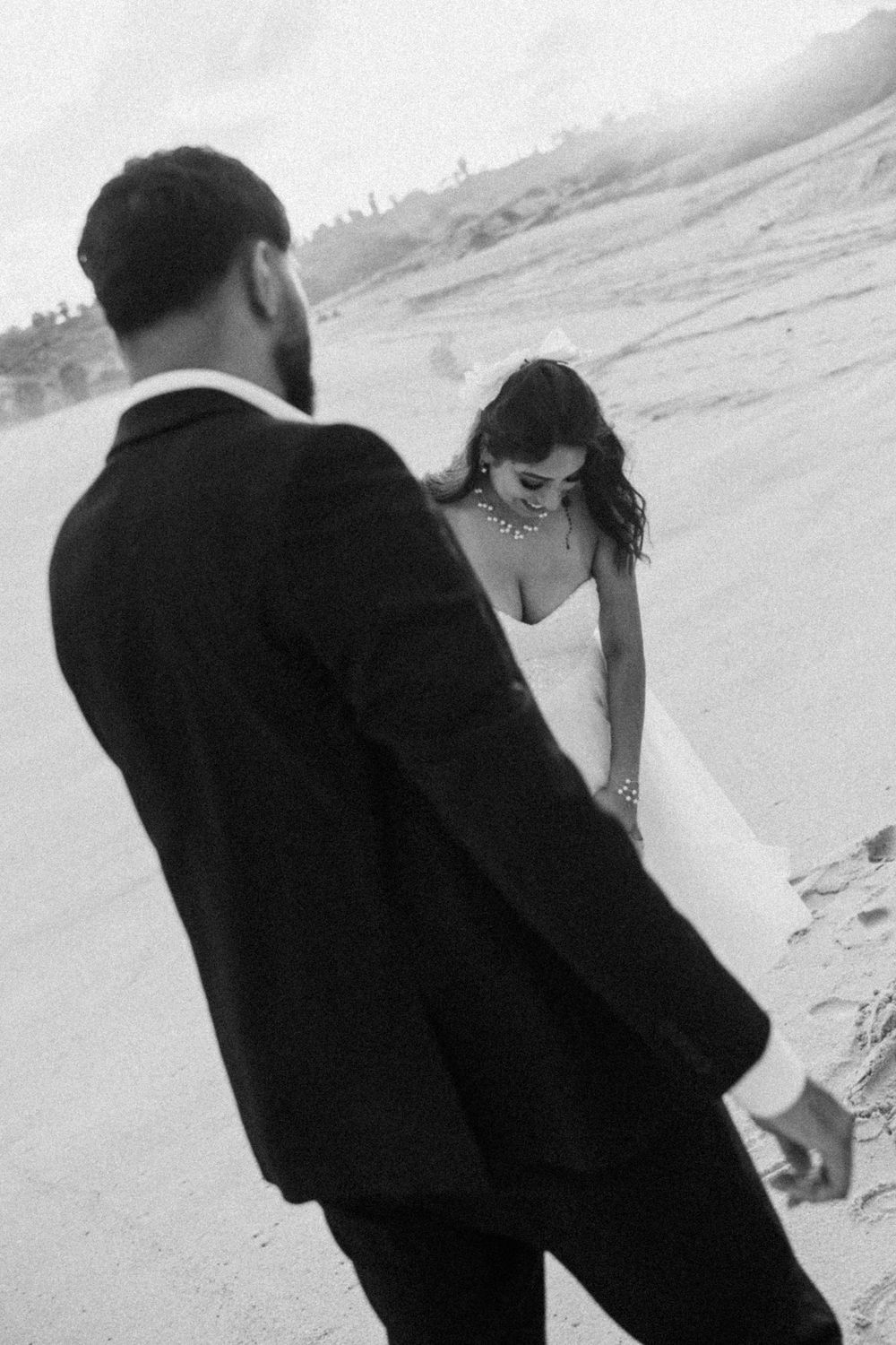 Black and white photo of a bride in white gown and groom in suit walking on a sandy beach.