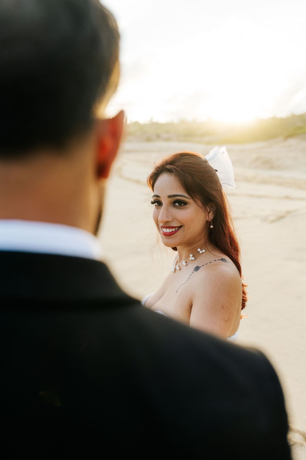 Bride in white gown and bow hair accessory smiles over shoulder at groom in black suit during golden hour beach wedding.