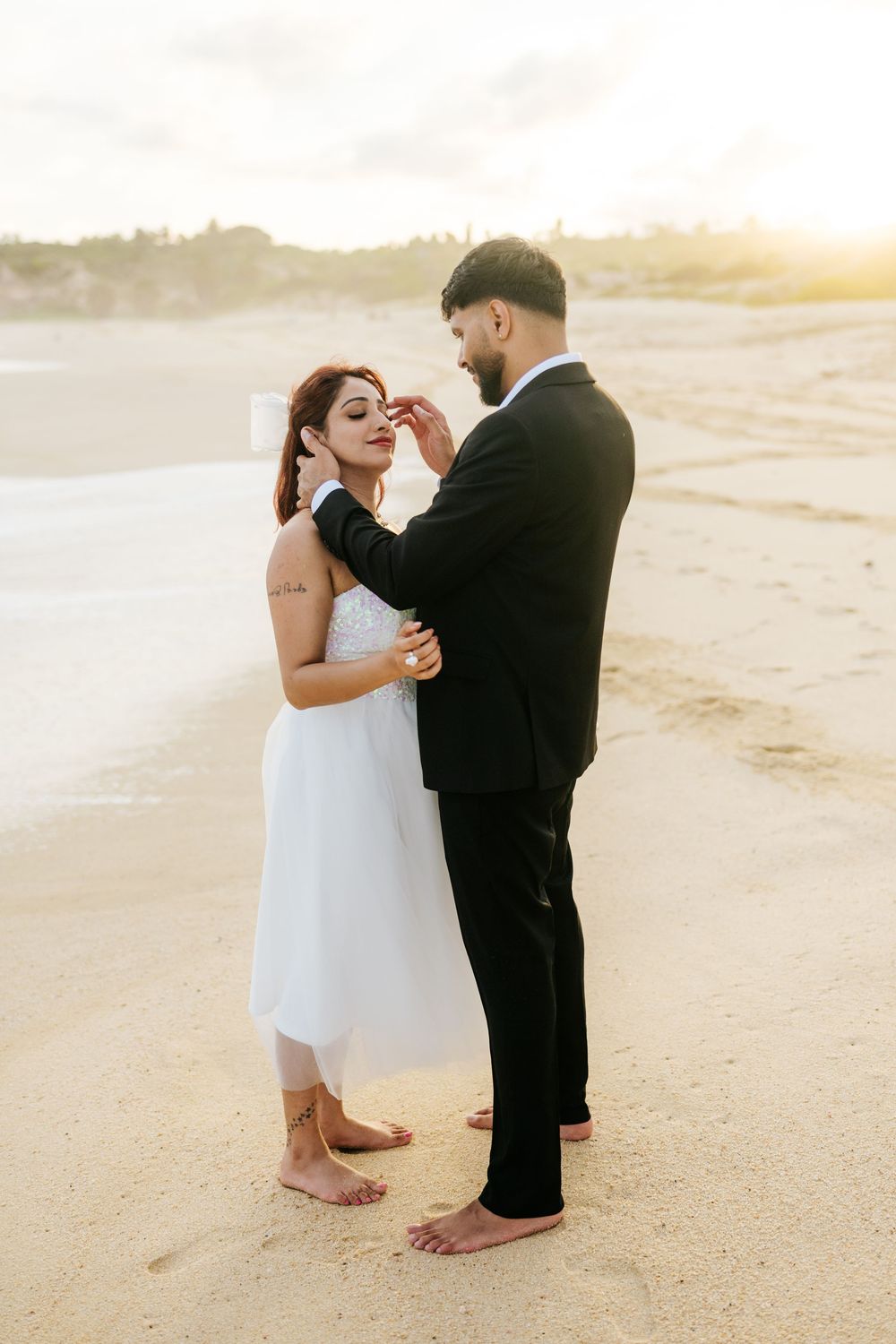 Couple in wedding attire sharing a romantic moment on a sandy beach at golden sunset, bride in white dress, groom in black suit.