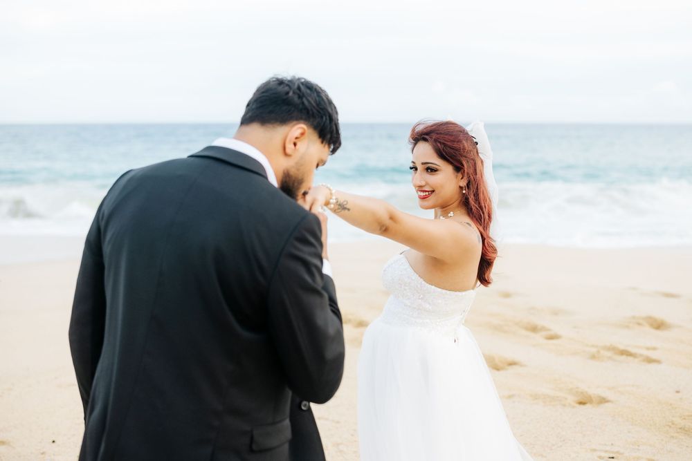 Bride and groom share a romantic moment on a sandy beach, with the groom kissing her hand by the ocean.