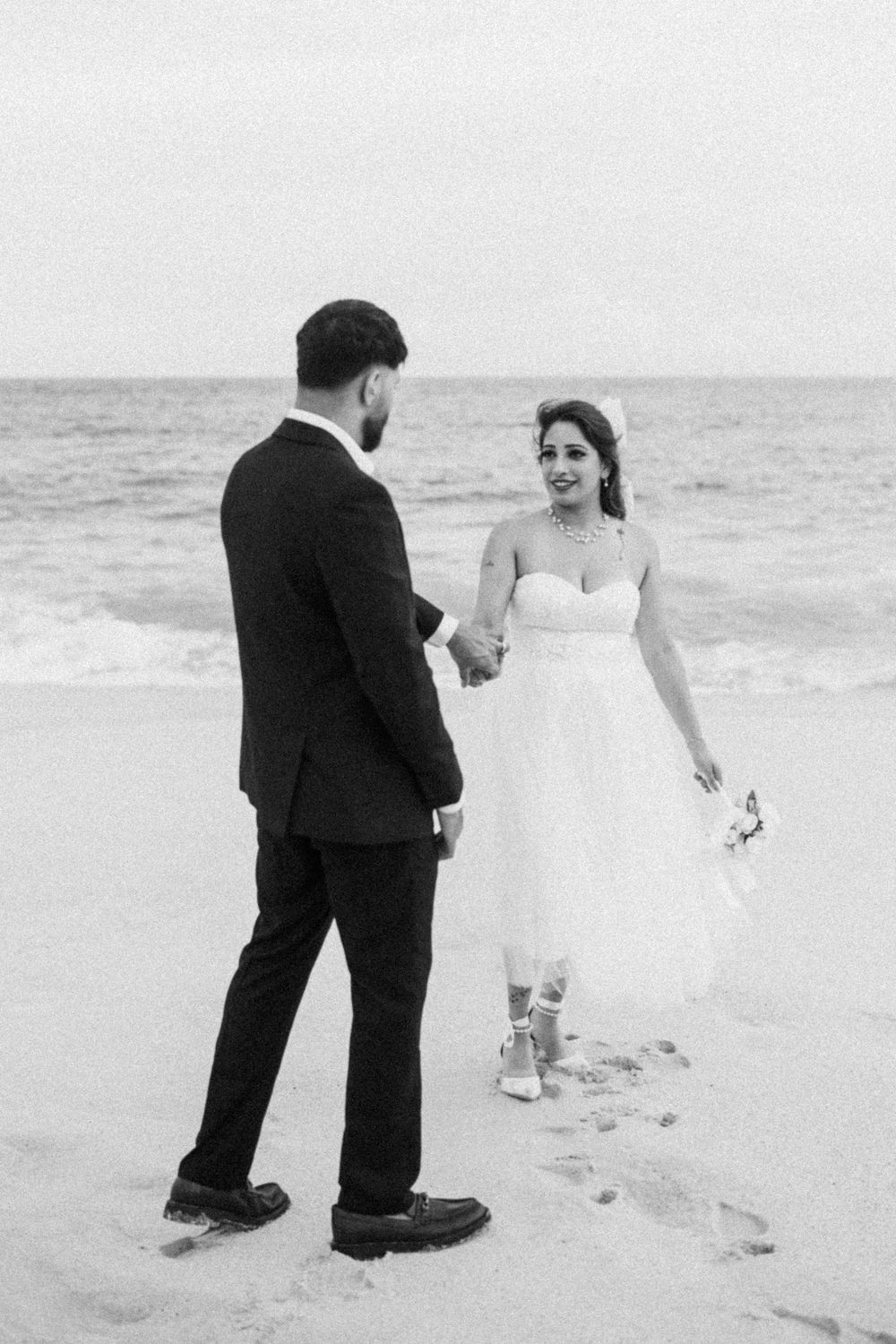 Black and white photo of a bride and groom holding hands on a beach, bride in short tulle dress smiling at groom.