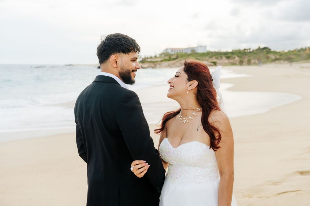 Bride and groom holding hands on a beach, smiling at each other, wearing wedding attire with ocean waves in the background.