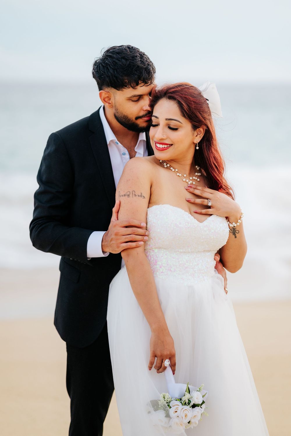 Couple in wedding attire embracing on a beach, bride in white strapless gown holding bouquet, groom in black suit.