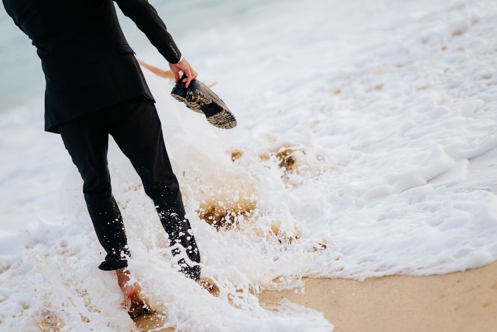 Person in black wetsuit wading through ocean waves on sandy beach while holding shoes.