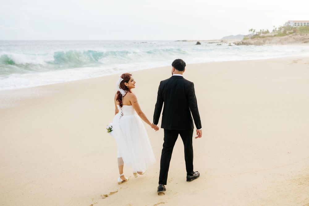 Bride and groom holding hands walking on a sandy beach with ocean waves in the background on their wedding day.
