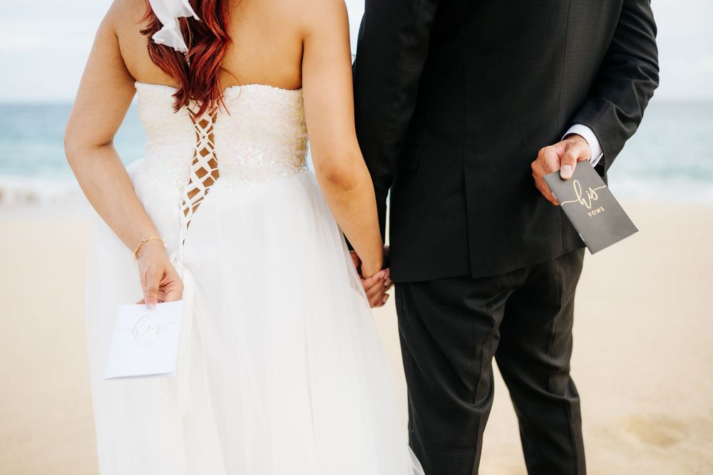 Bride in strapless lace wedding gown and groom in black suit holding hands on a sandy beach.