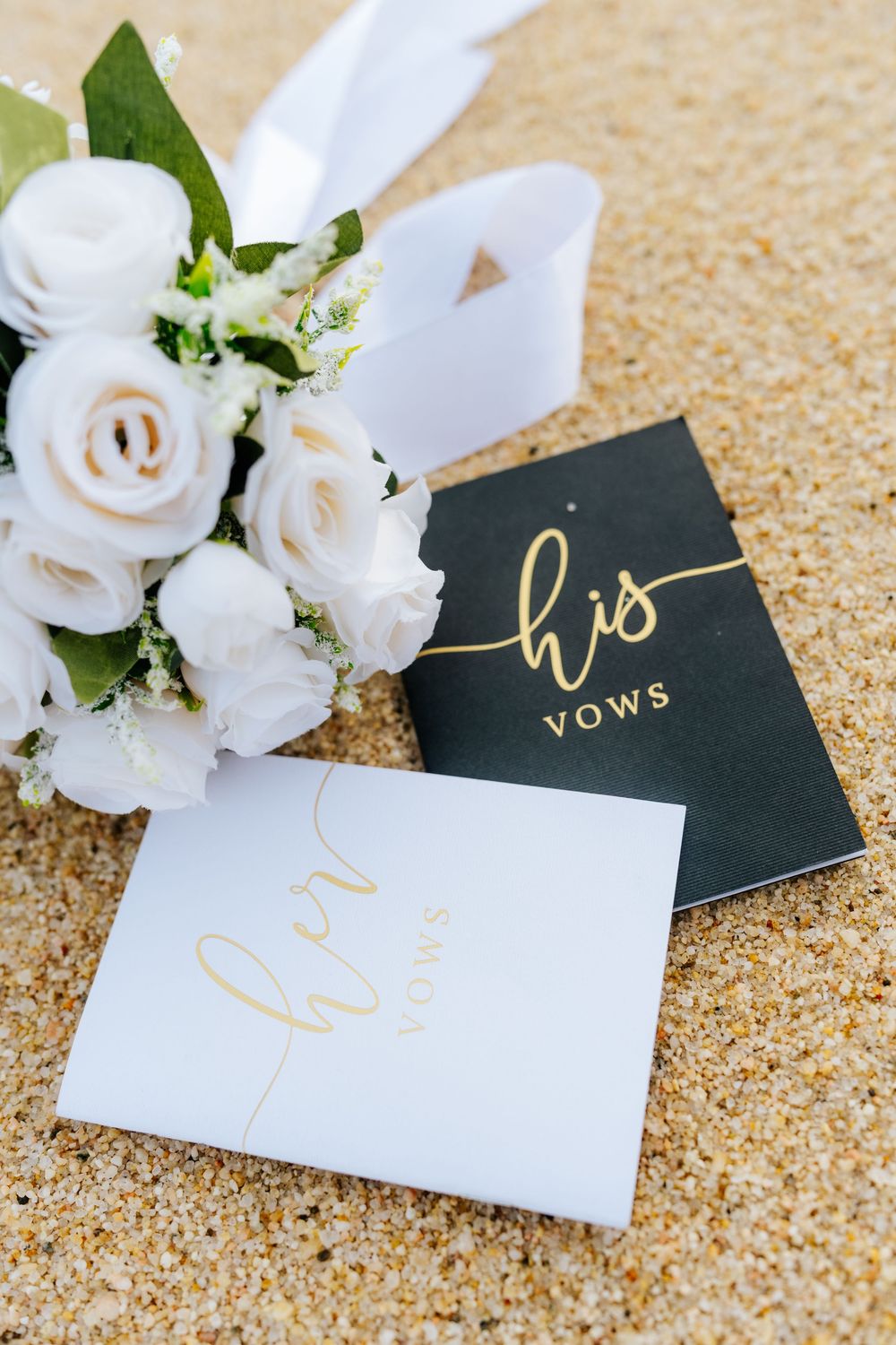 His and her wedding vow books in black and white with gold script, beside a white rose bouquet on sandy beach.