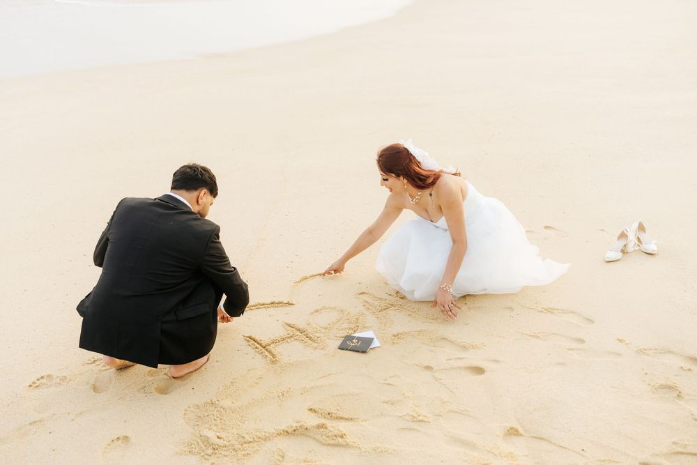Bride and groom writing in sand on beach, woman in white dress and man in black suit crouching together.