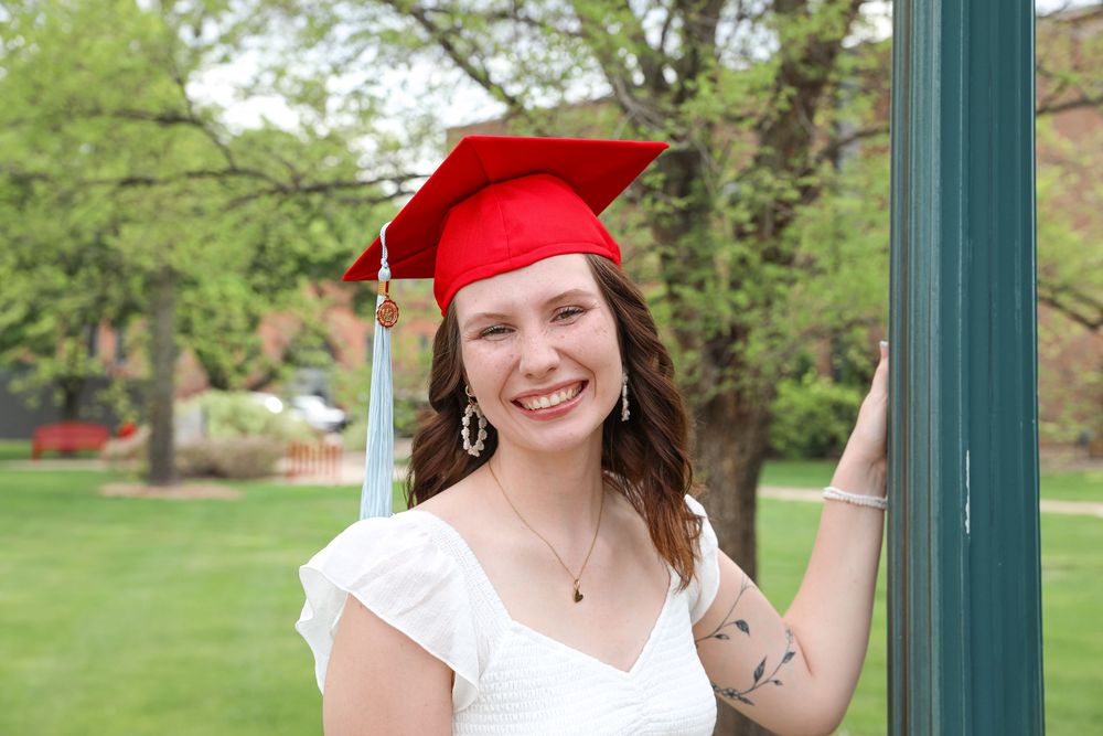 Smiling young woman in red graduation cap and white dress posing outdoors on a sunny day.