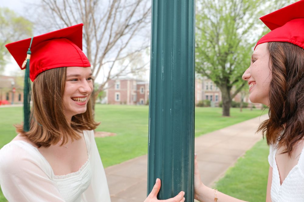 Two smiling female graduates in red caps stand by a green pole on a college campus.