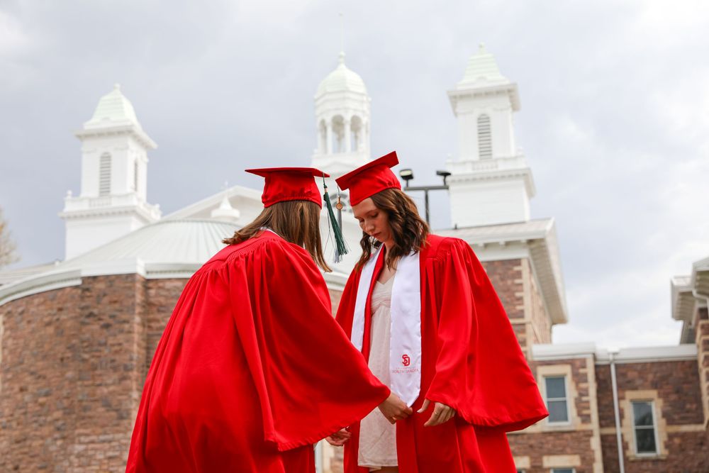 Two graduates in red caps and gowns hold hands outside a historic brick building on graduation day.