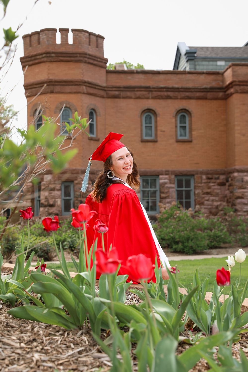 Smiling graduate in red cap and gown stands among red tulips in front of a brick building.