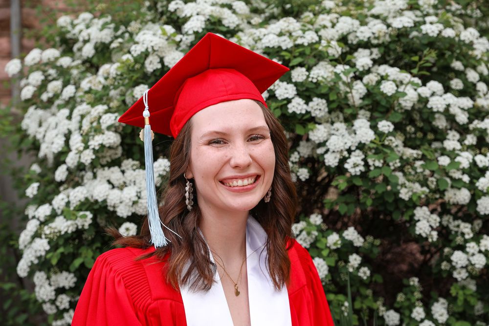 Smiling graduate in red cap and gown poses in front of white flowering bushes at outdoor graduation ceremony.