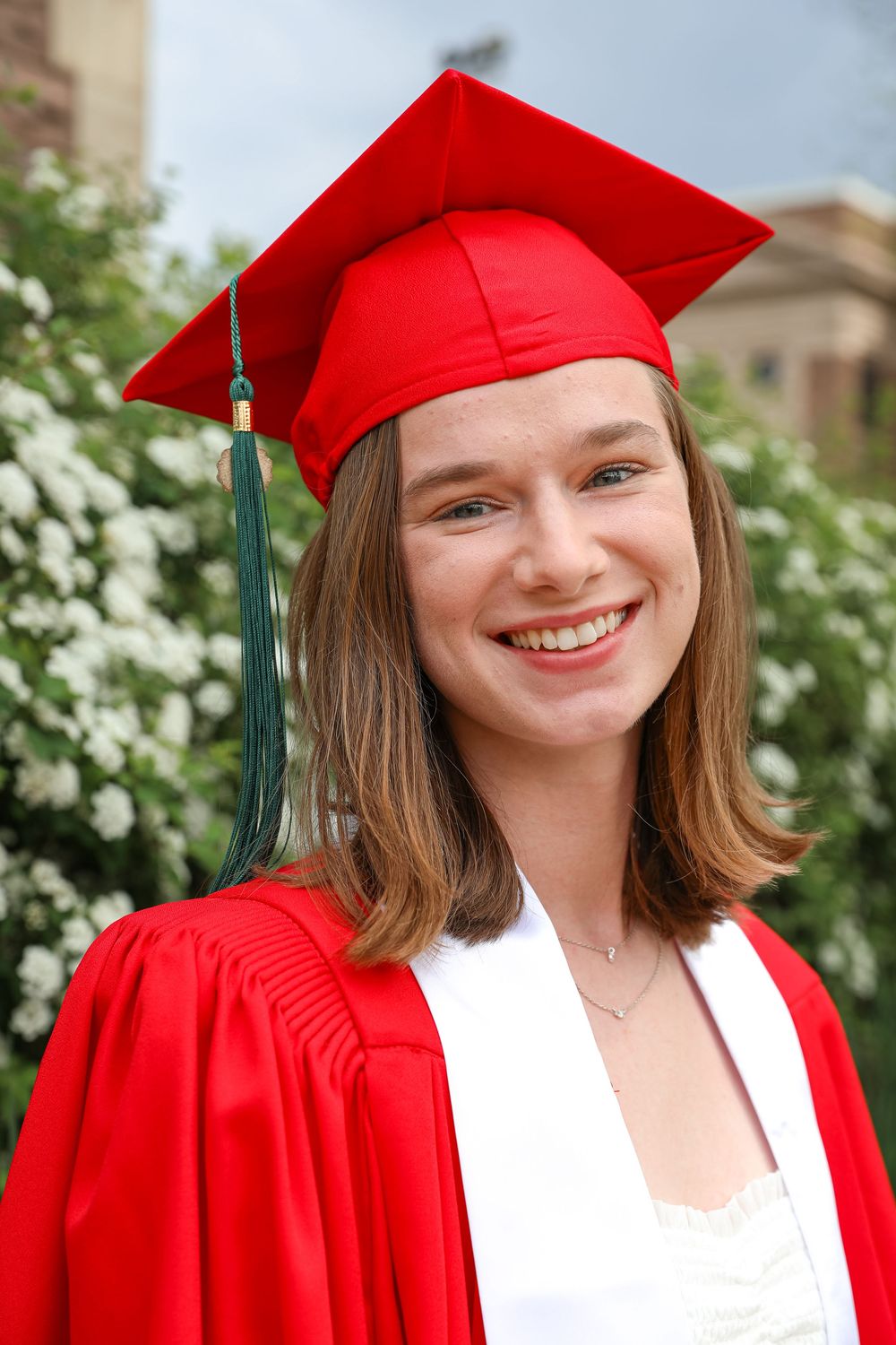 Smiling young woman in red graduation cap and gown posing outdoors with white flowering bushes in background.