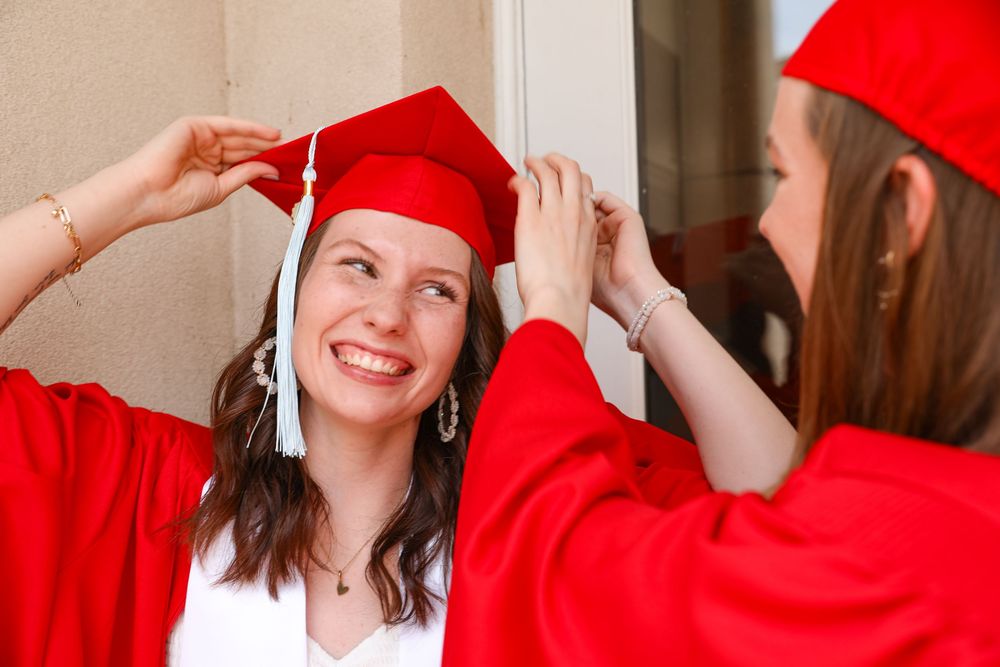 Two young women in red graduation caps and gowns smiling as one adjusts the others mortarboard cap.