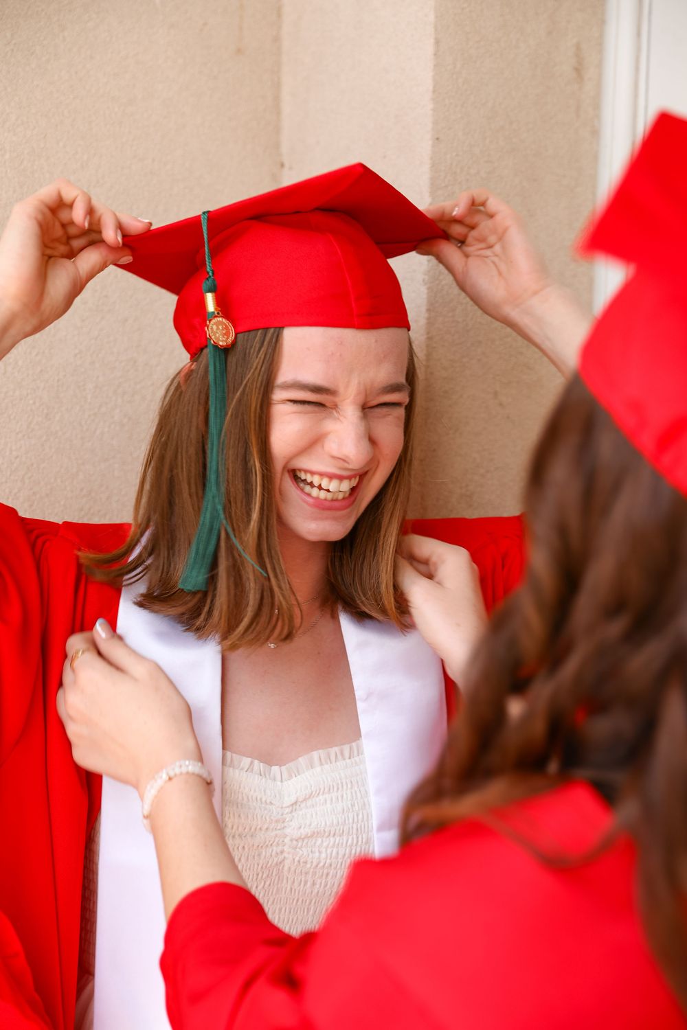 Laughing girl in red graduation cap and gown being helped by friend outdoors.