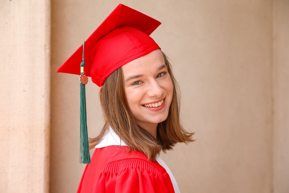 Smiling young woman in red graduation cap and gown celebrating academic achievement outdoors.