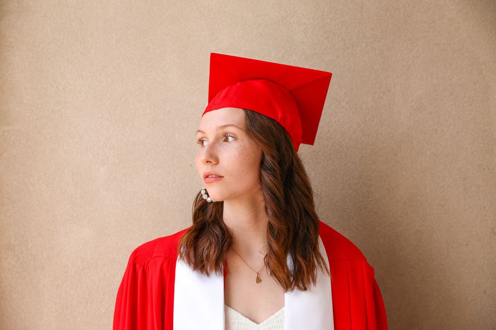 Young woman wearing red graduation cap and gown, looking upward thoughtfully against a neutral beige background.