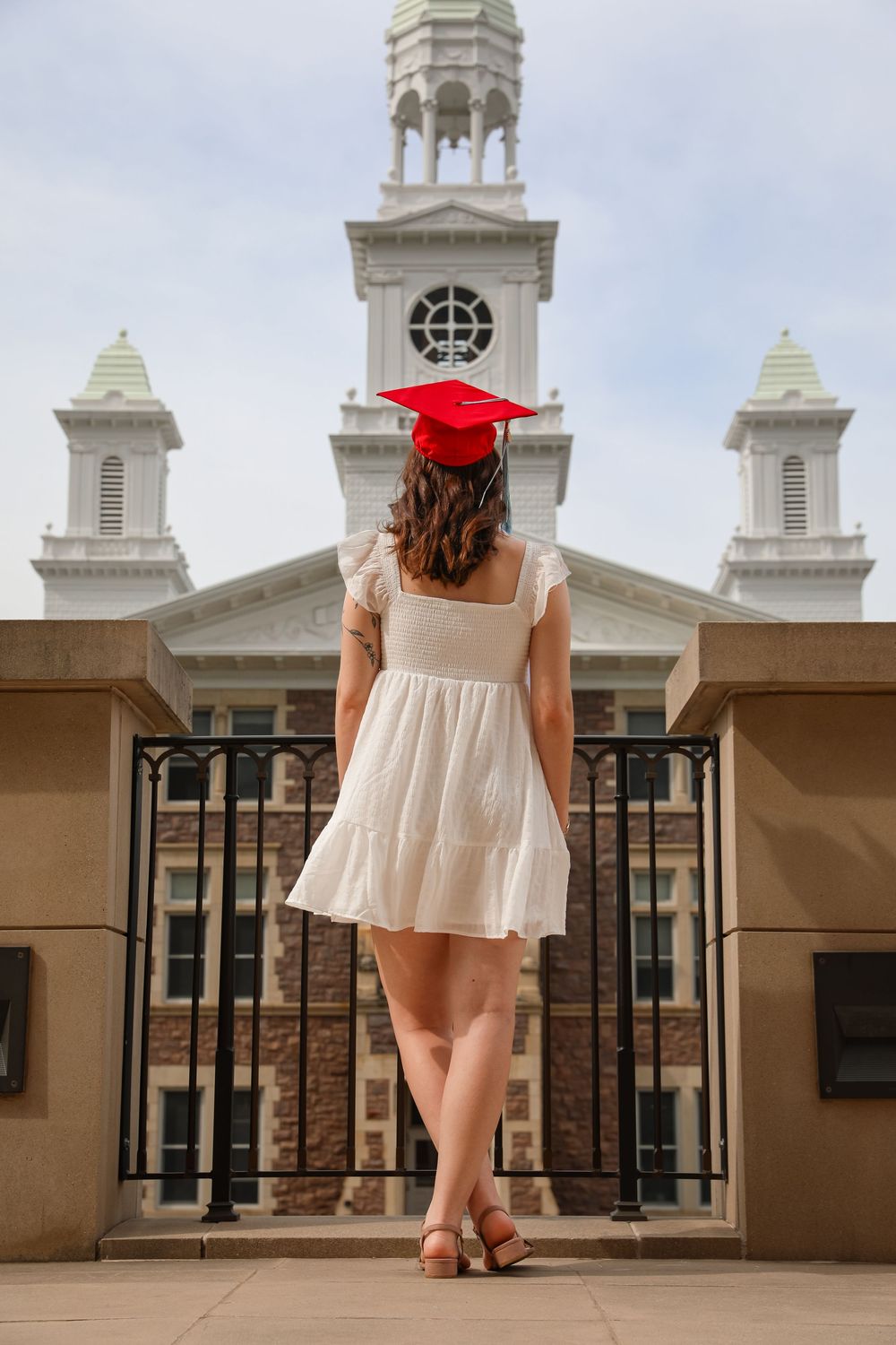 Young woman in white dress and red graduation cap stands before a classic university building with white towers.