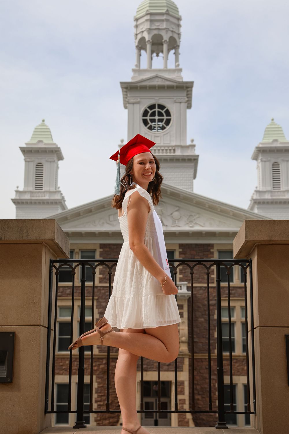 Young woman in white dress and red graduation cap poses joyfully in front of a classic brick university building.