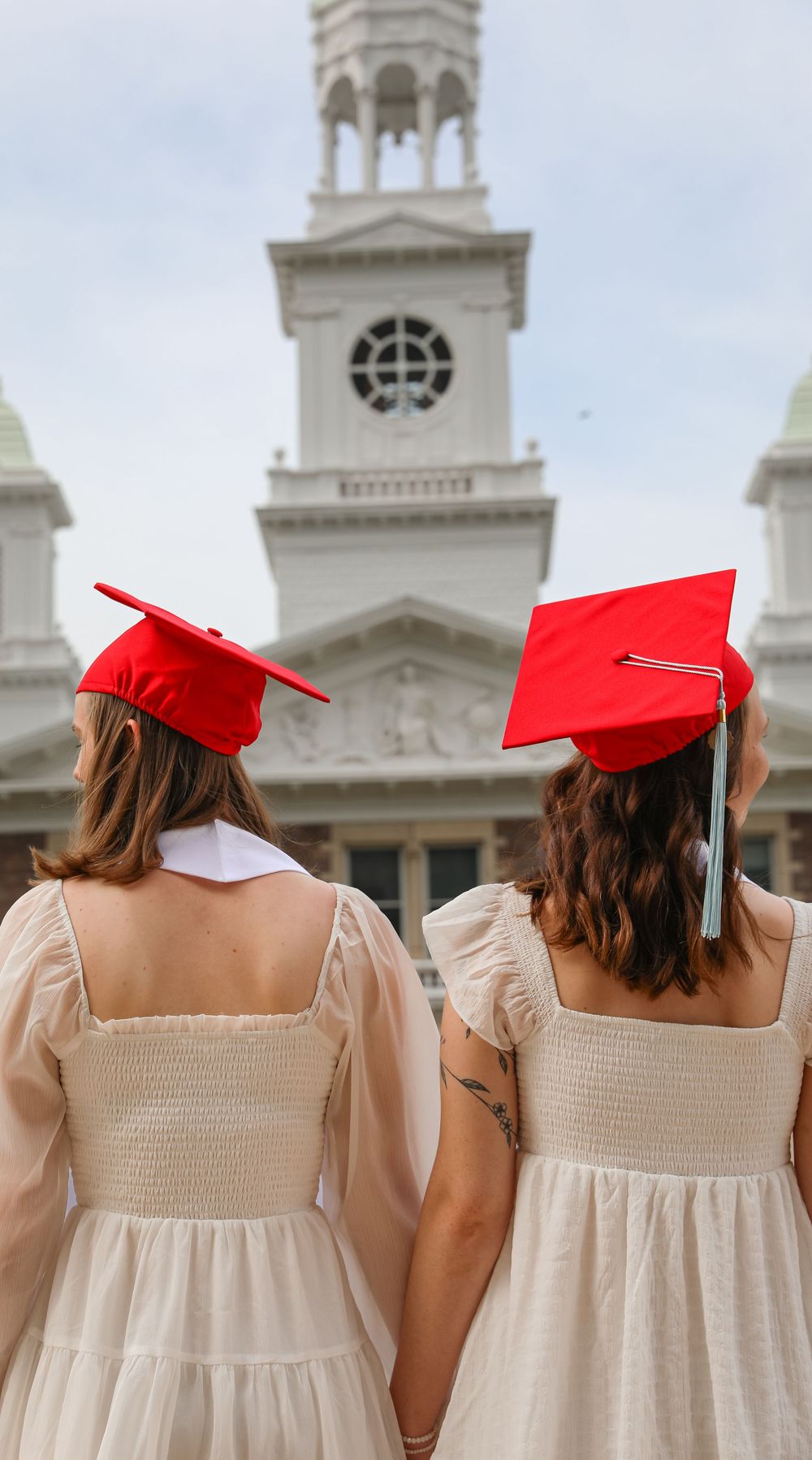 Two graduates in red caps and cream dresses hold hands facing a white neoclassical building.