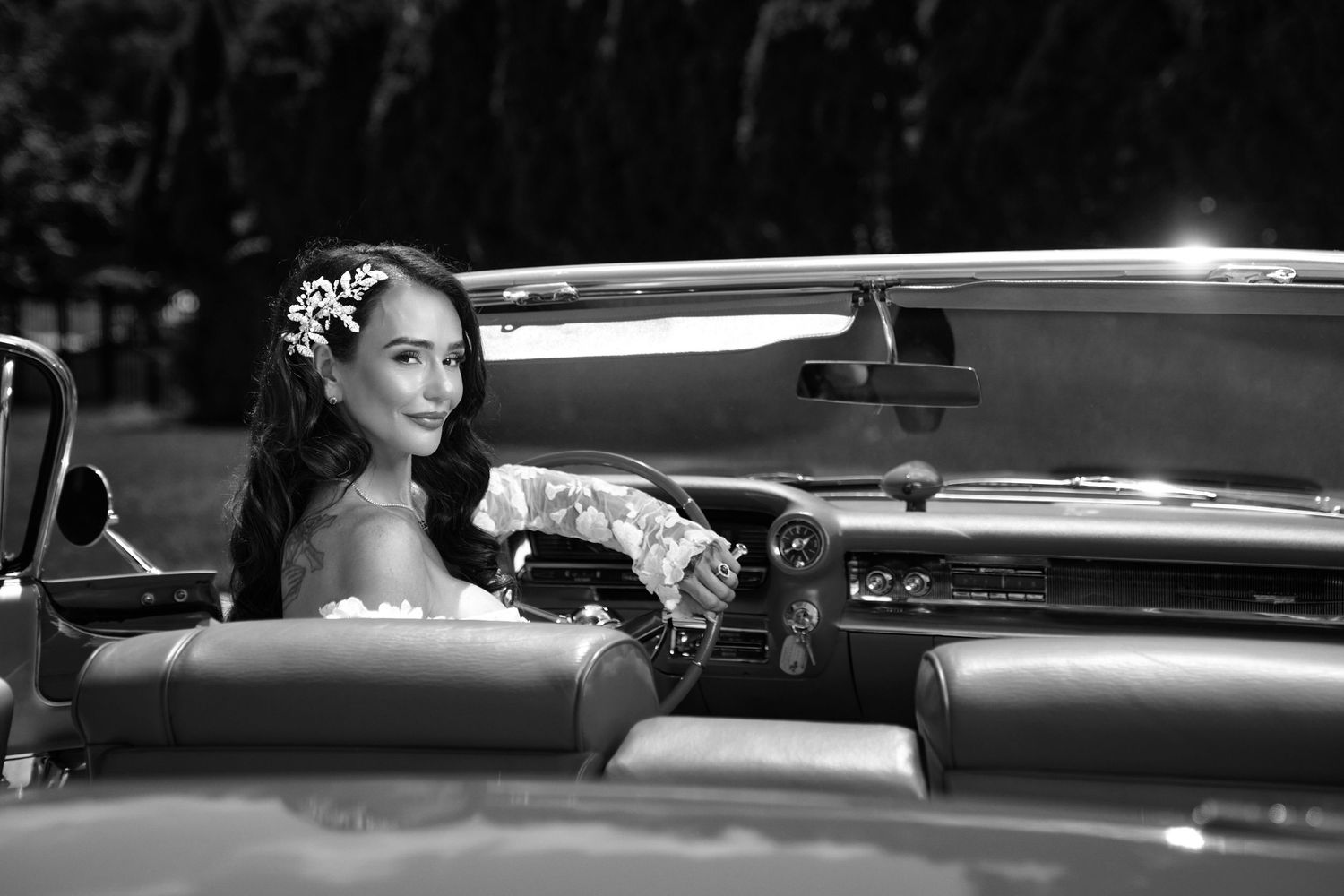 Bride in floral headpiece sits elegantly in a classic convertible car, captured in stunning black and white photography.