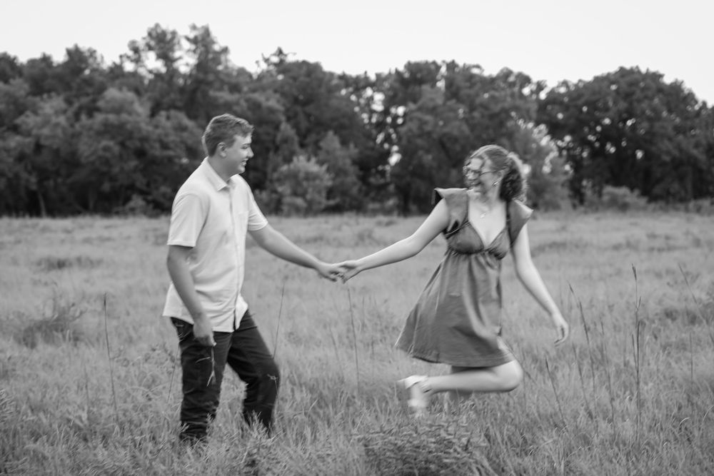 Black and white photo of a couple holding hands in a grassy field, woman jumping joyfully.