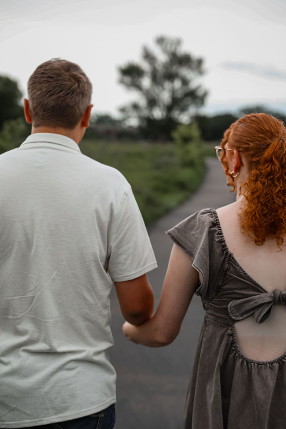 Couple holding hands walking down a path surrounded by greenery, viewed from behind.