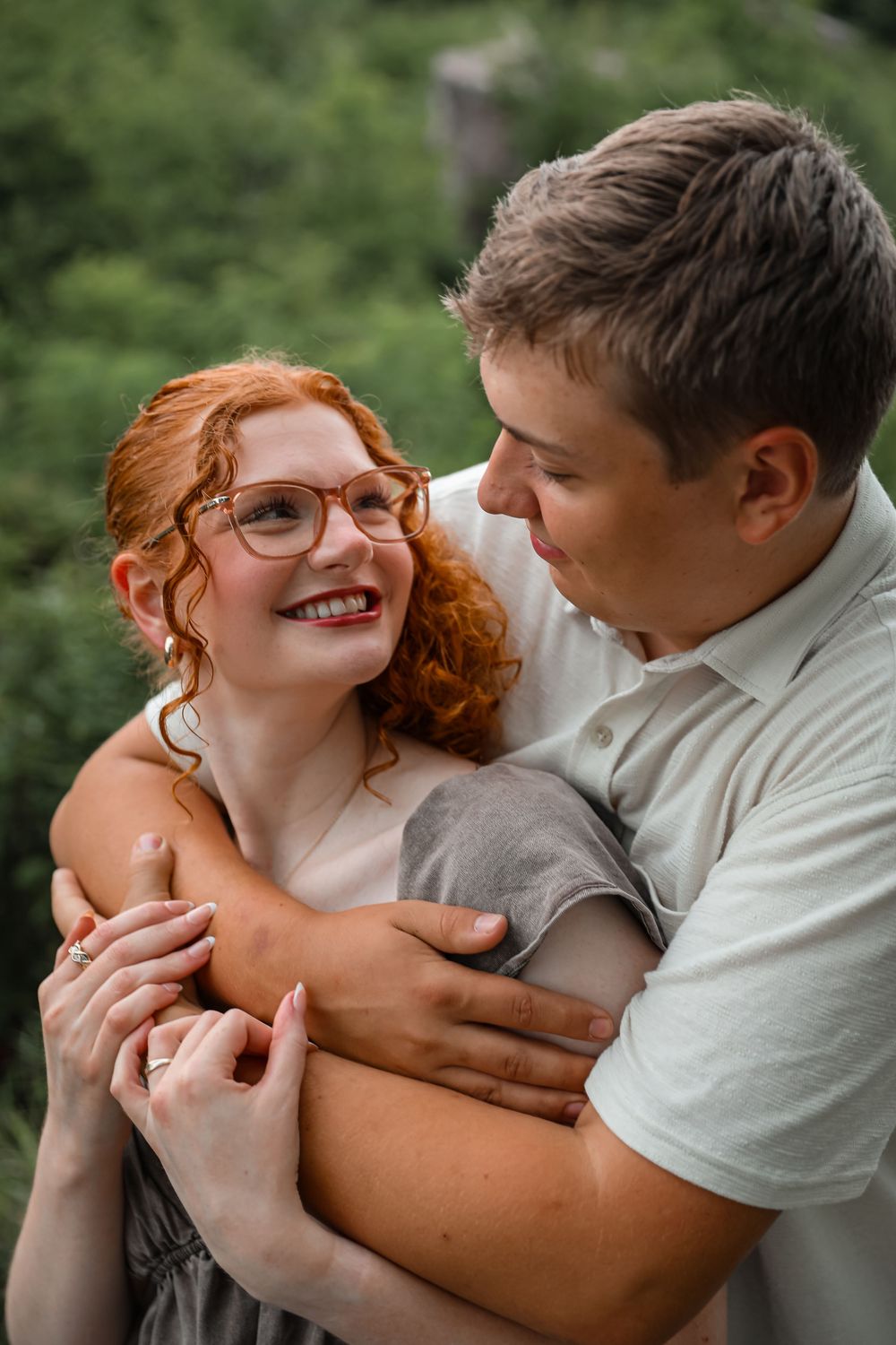 A smiling redhead woman with glasses embraces a man outdoors, surrounded by lush green foliage.