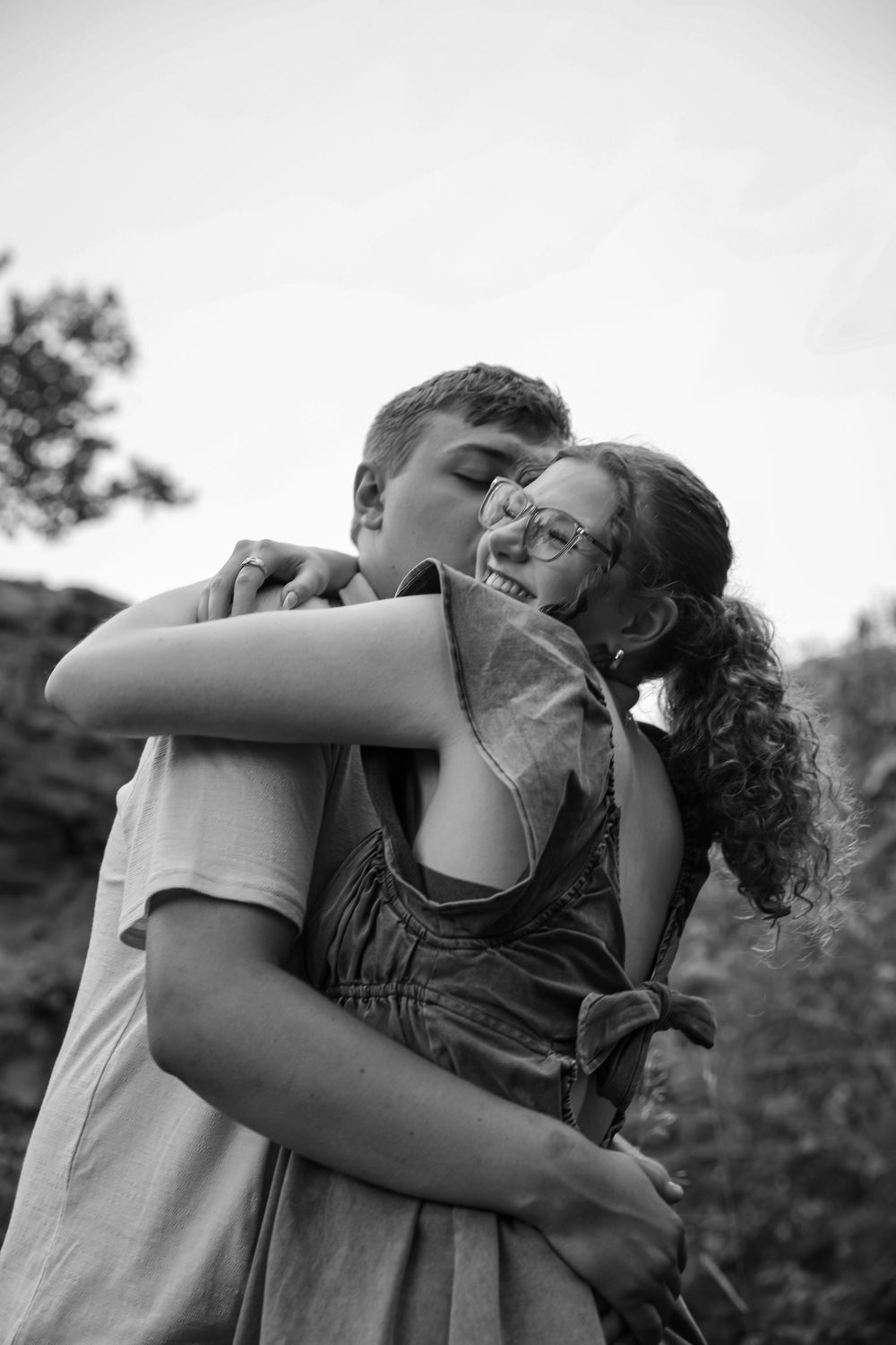 Black and white photo of a young couple embracing outdoors, man kissing woman's cheek as she smiles warmly.