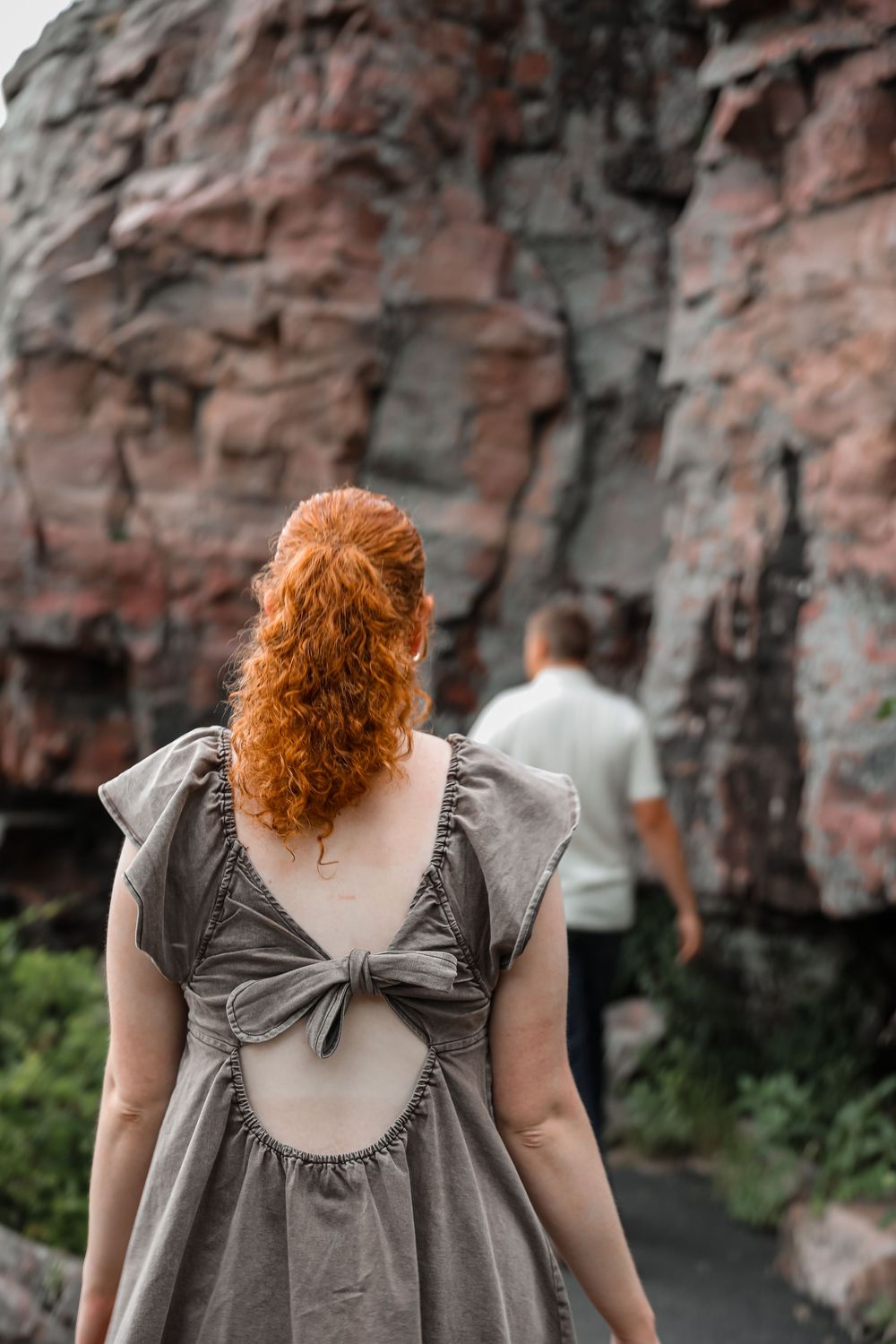 Red-haired woman in gray open-back bow dress stands before rocky cliff with man in background.