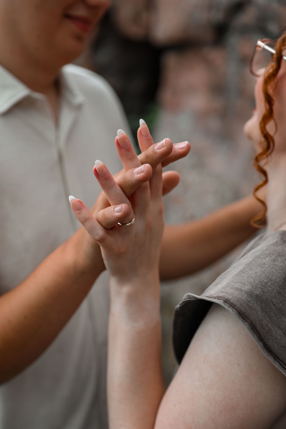 Close-up of a couple holding hands, woman wearing a gold ring with French manicure nails outdoors.