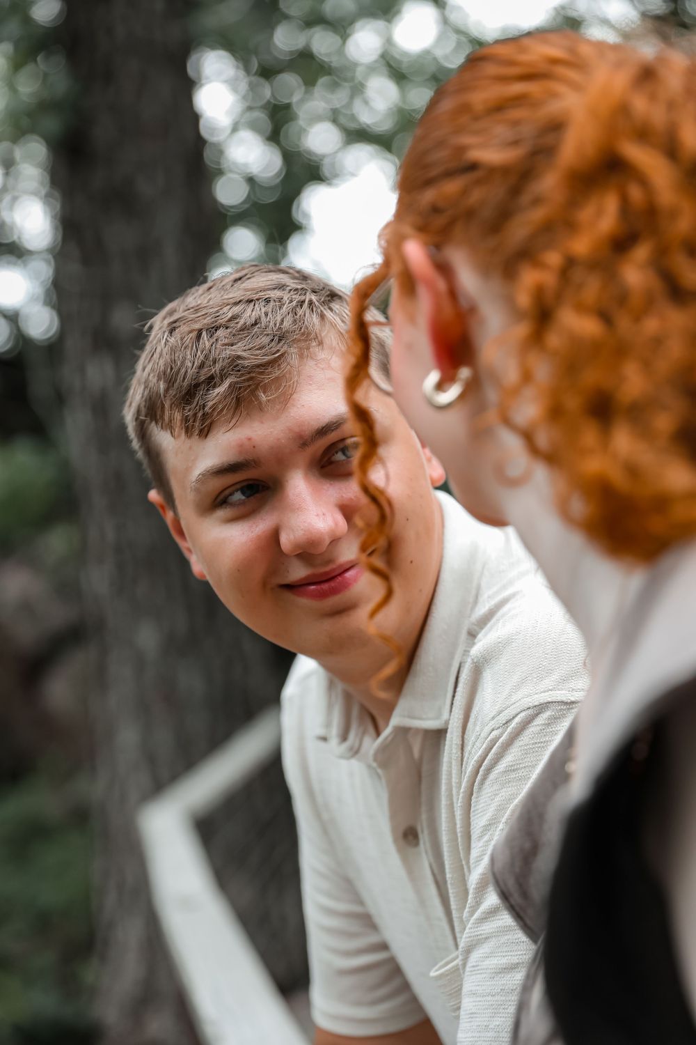 A young man in a white polo shirt smiles warmly at a redheaded person in an outdoor setting.