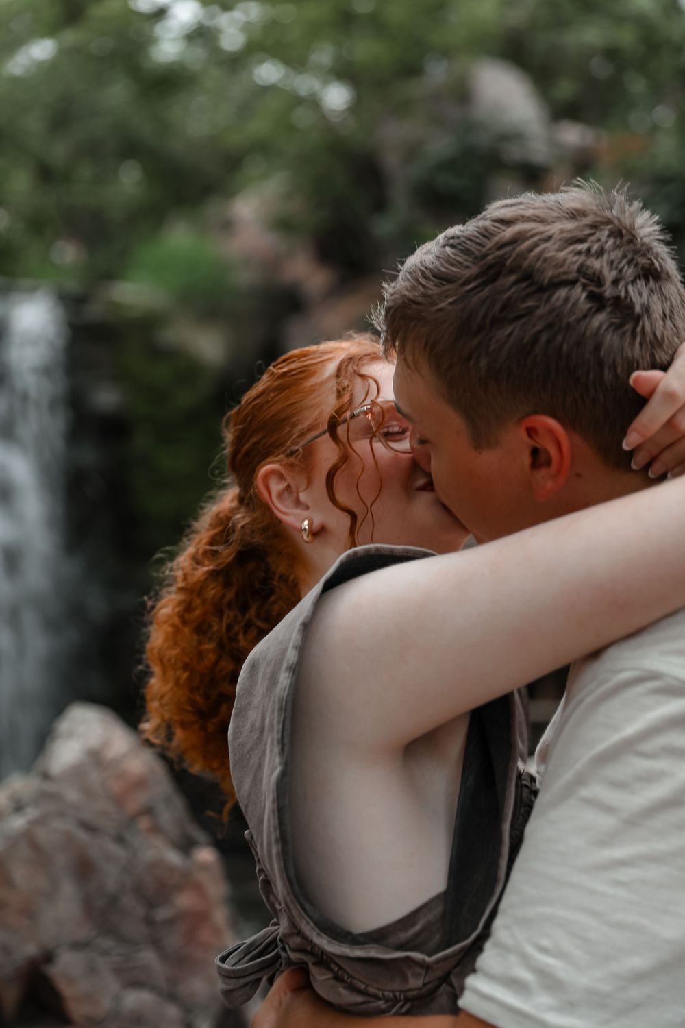 A couple shares a romantic kiss near a waterfall, the woman with red hair embracing the man outdoors.