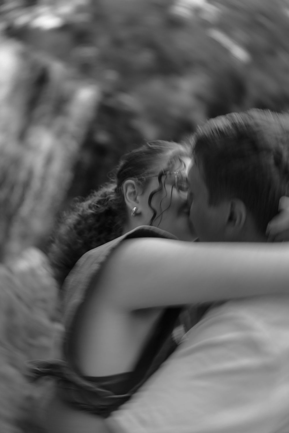 Black and white photo of two people sharing an intimate embrace outdoors with blurred natural background.