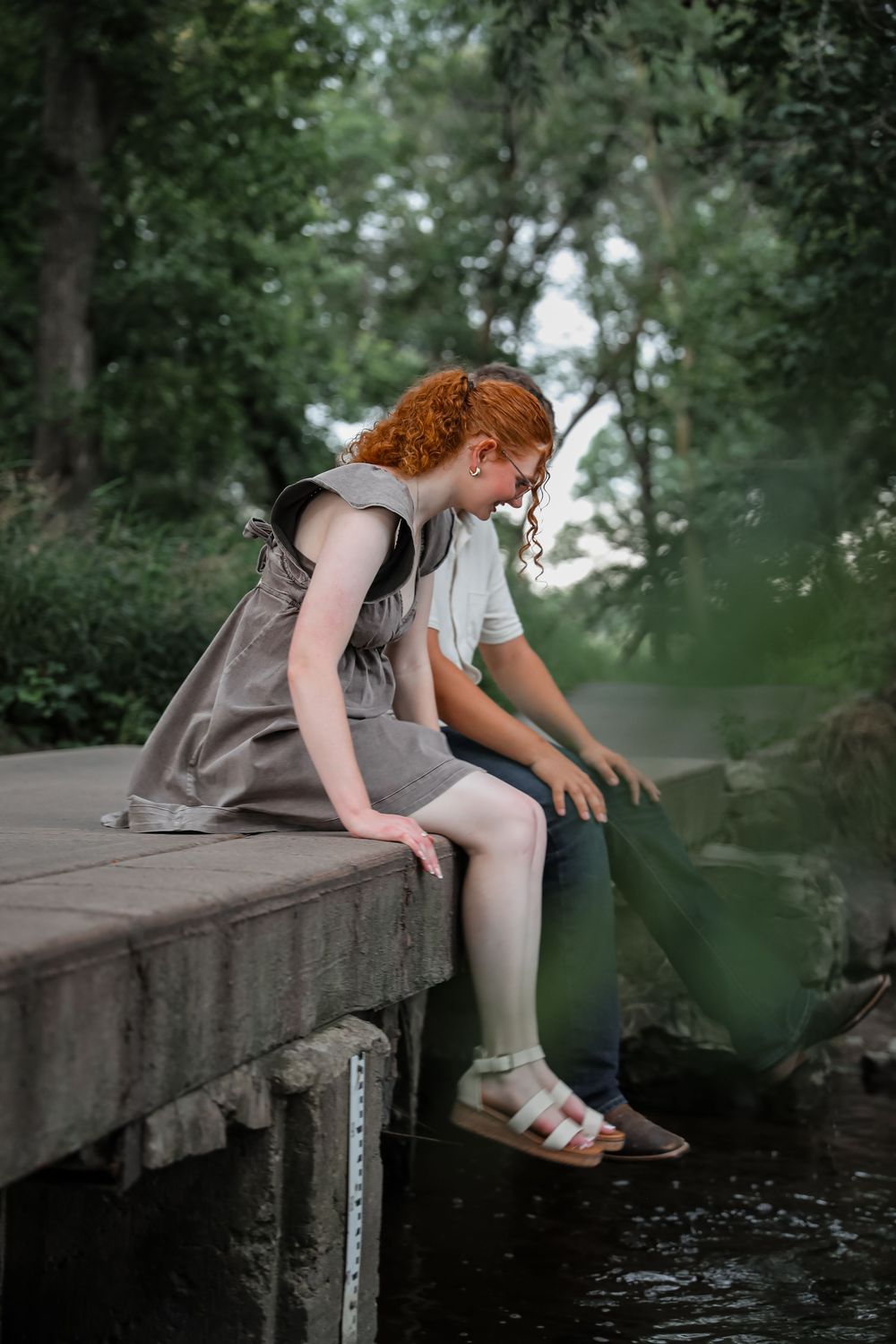 Red-haired girl in gray dress sitting on a concrete bridge edge near a creek, looking down at water.
